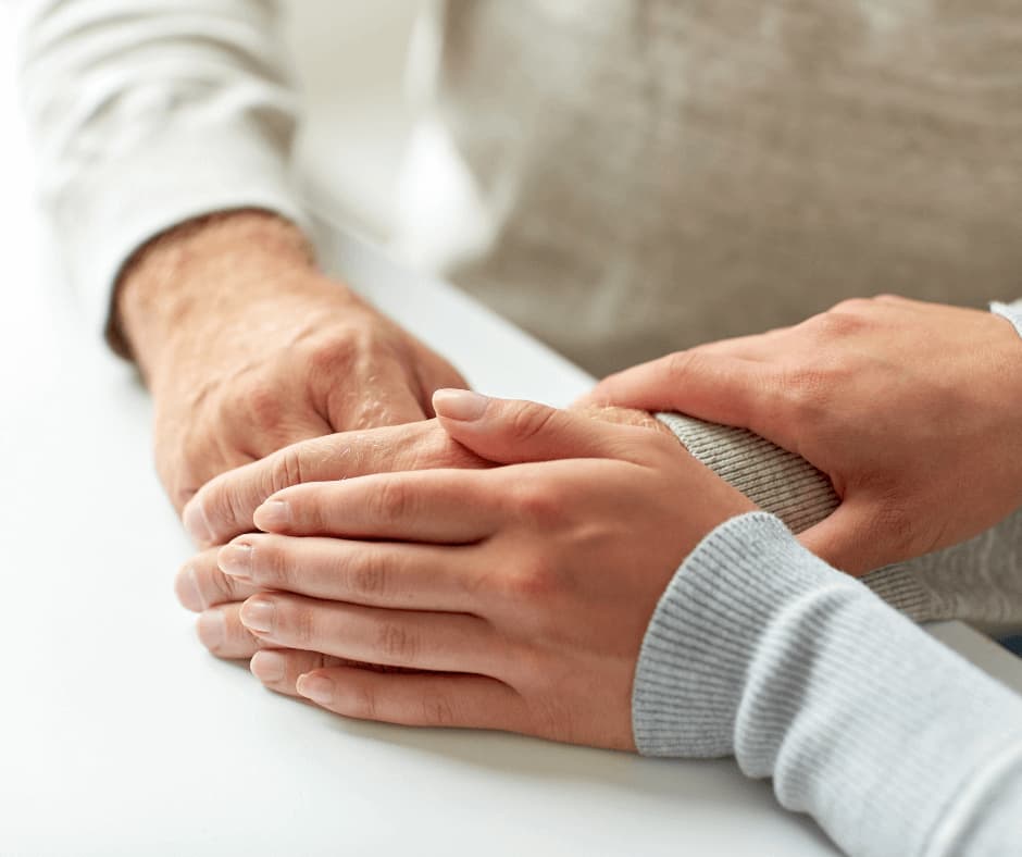 A close-up of two people holding hands, offering comfort and support to each other across a white table. - Home Instead