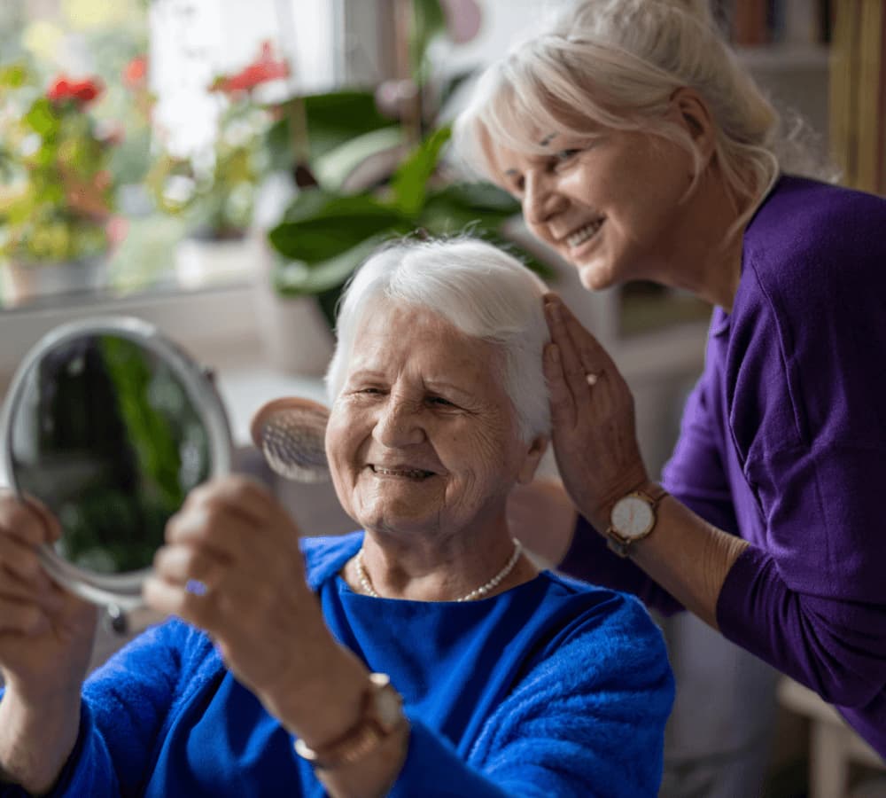 Two elderly women smiling, with one holding a mirror while the other gently touches her shoulder in a brightly lit room. - Home Instead