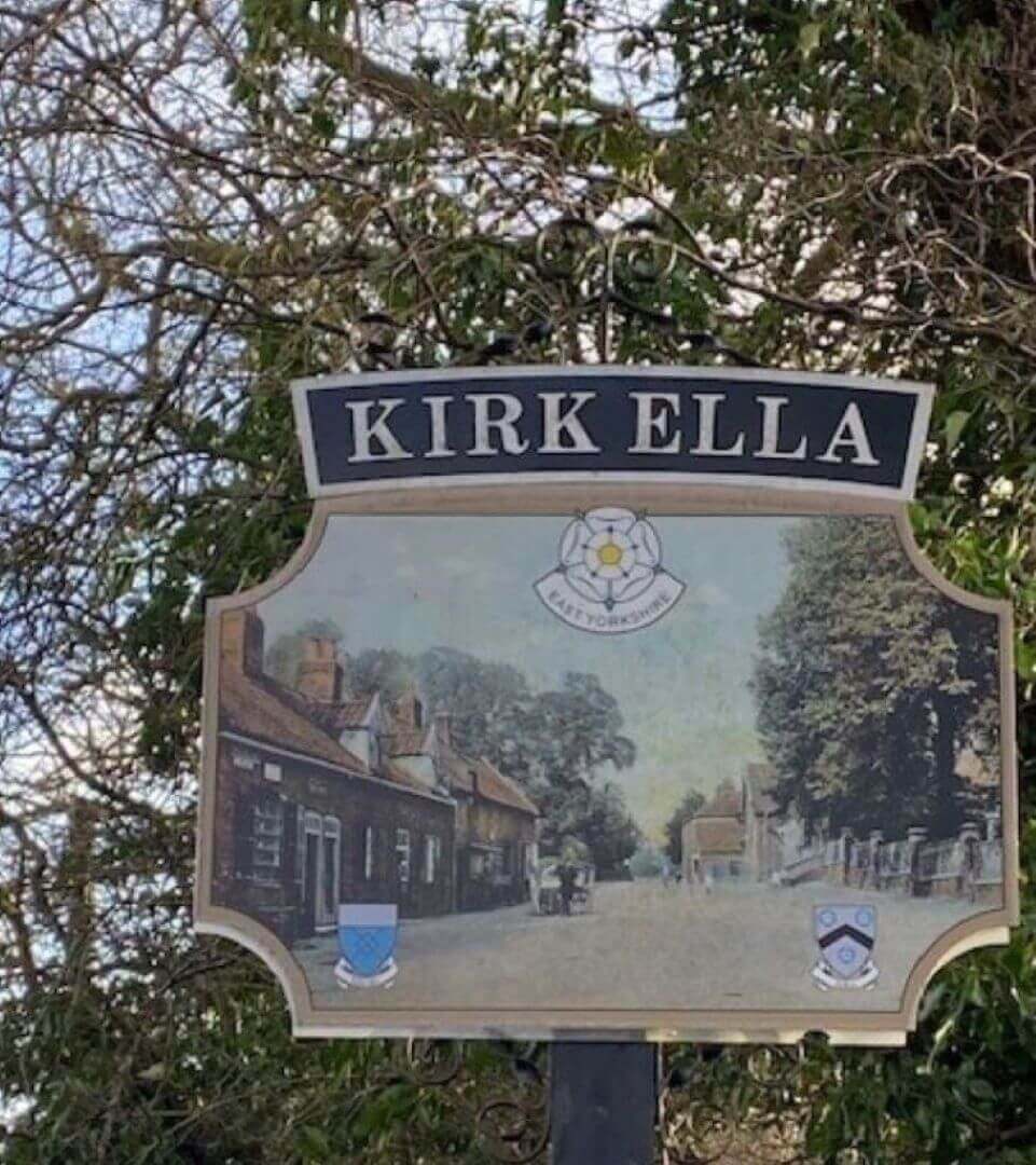 Kirk Ella sign with Yorkshire emblem and a historic street scene in front of trees and a cloudy sky. - Home Instead