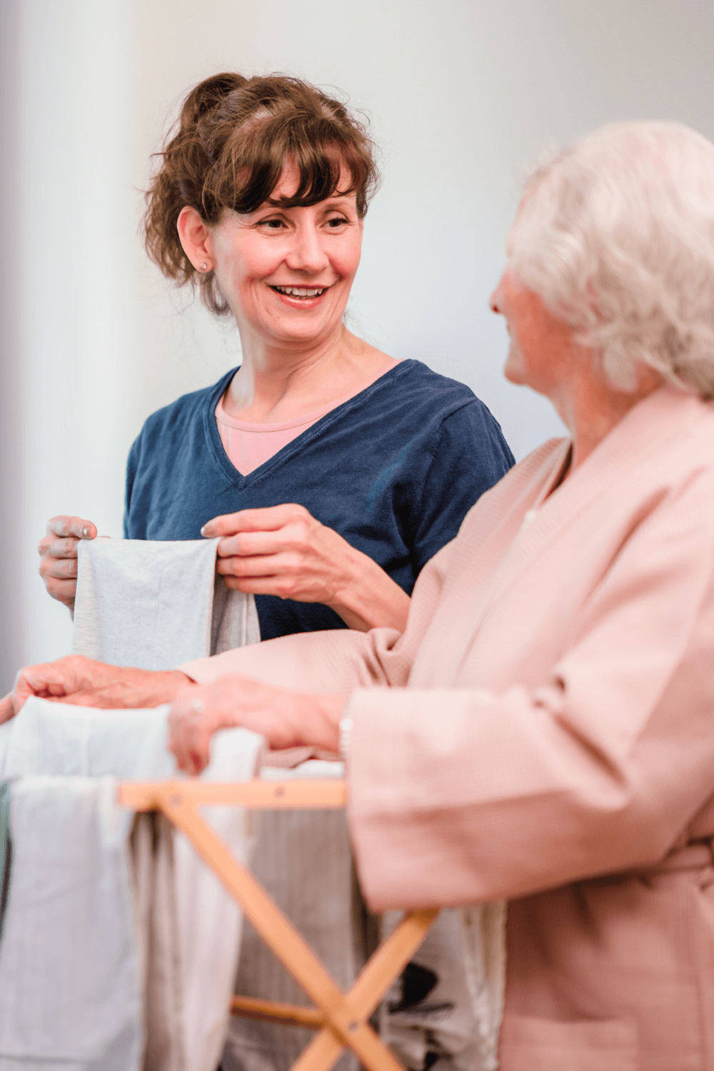Two women smiling while folding laundry together, one in a blue shirt and the other in a light pink robe. - Home Instead