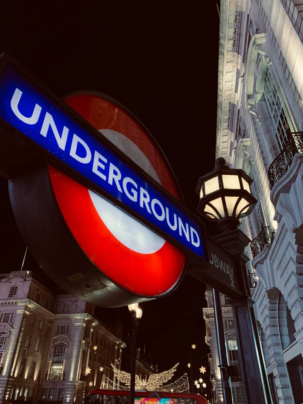 London Underground sign at night with illuminated buildings and decorative lights in the background. - Home Instead