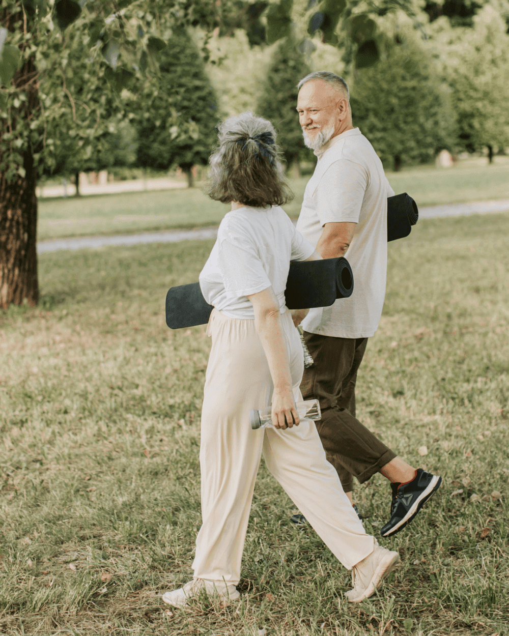 Two people walking in a park, each carrying a yoga mat, with trees and grass in the background. - Home Instead