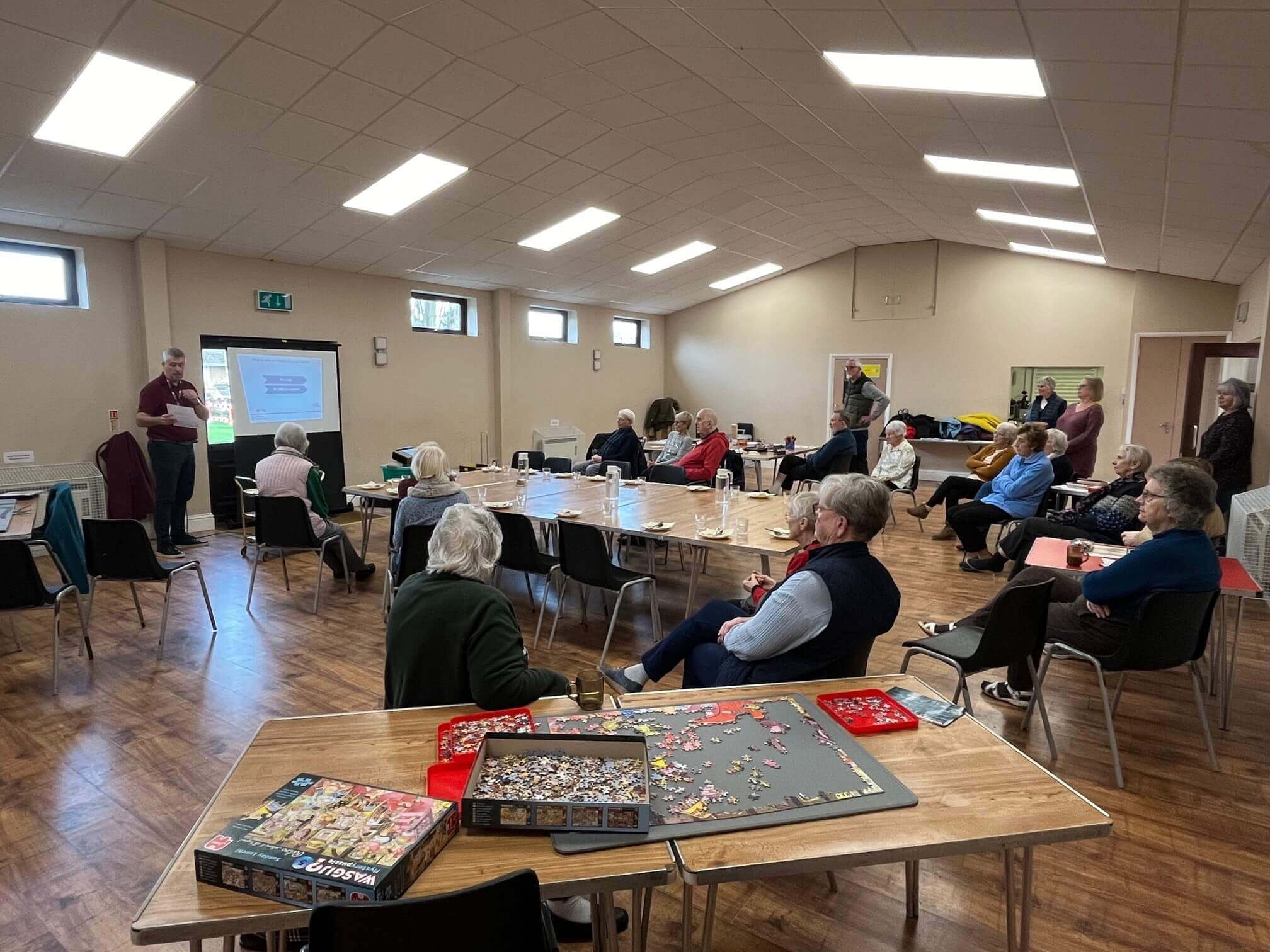 A group of elderly individuals gather in a community hall for a presentation, with puzzles on tables. - Home Instead