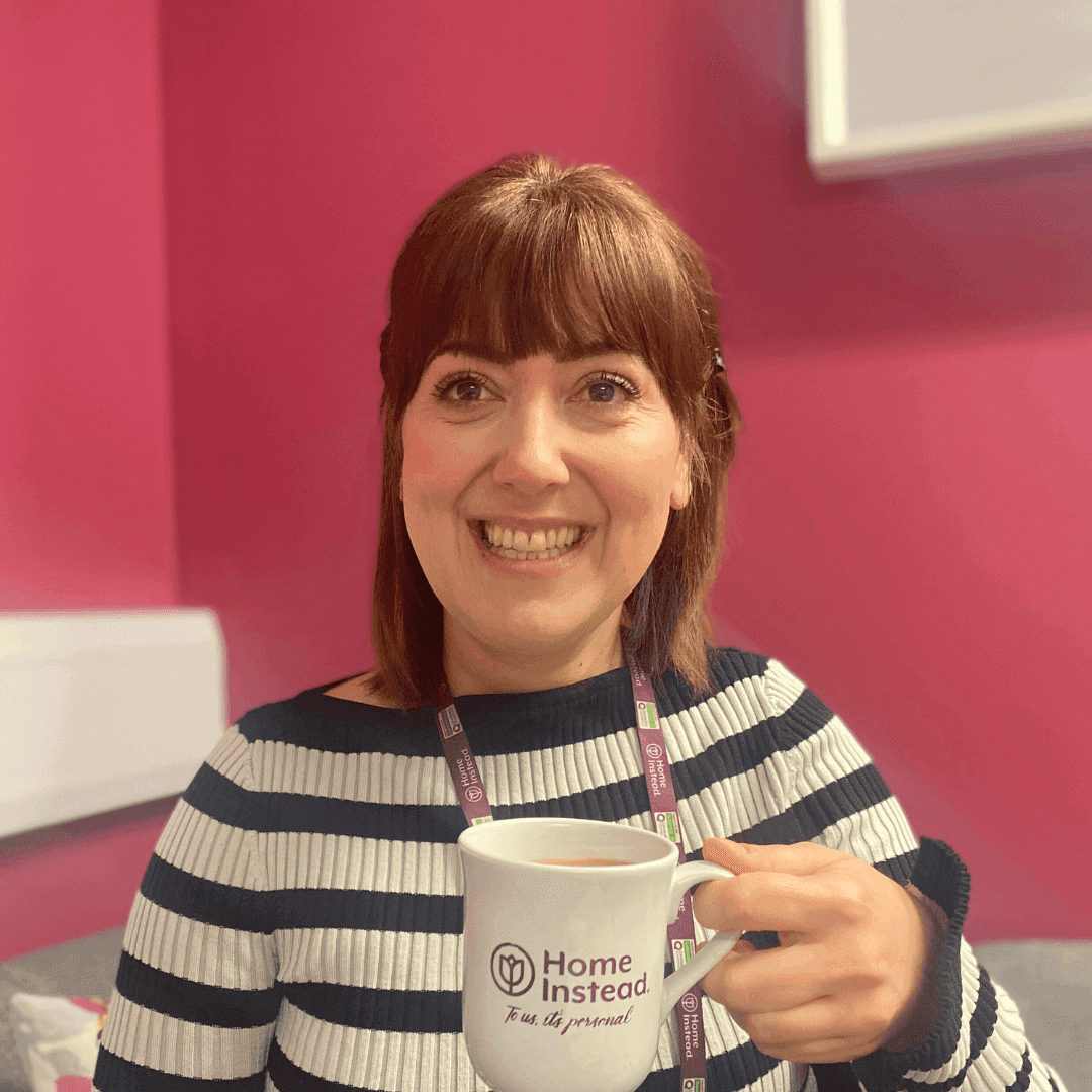 Smiling woman holds a "Home Instead" mug in front of a pink background, wearing a striped shirt and lanyard. - Home Instead