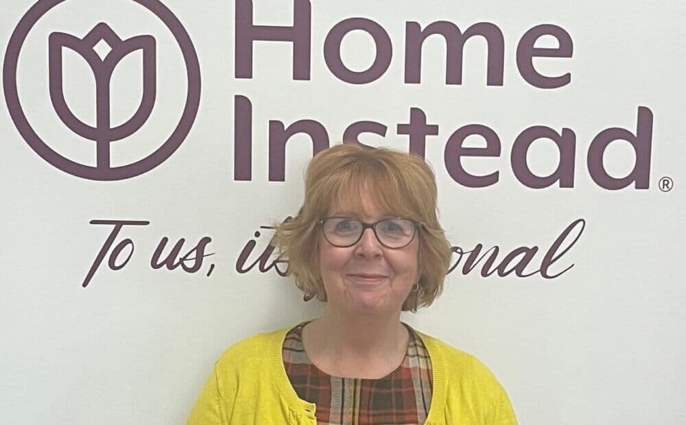 A woman with short hair stands smiling in front of a Home Instead logo on a wall. She is wearing glasses and a yellow cardigan. - Home Instead