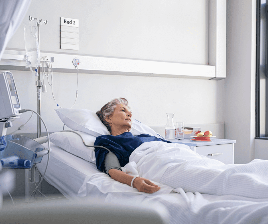 Elderly woman in hospital bed with medical equipment, IV drip, and glass of water on bedside table, sign above reads "Bed 2". - Home Instead