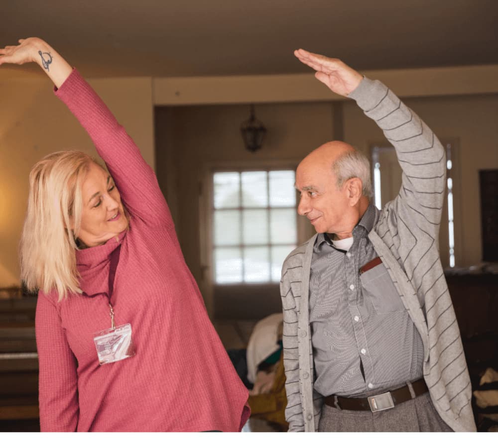 A smiling woman and an elderly man stretch their arms overhead in a well-lit room. - Home Instead