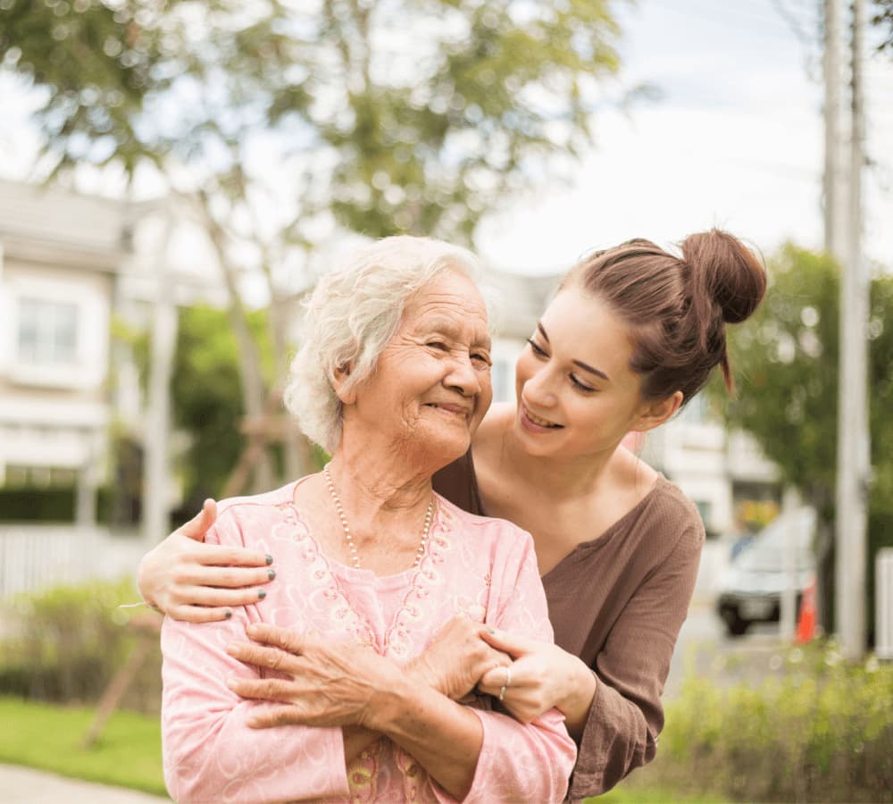 Young woman embraces elderly woman from behind while they smile at each other in an outdoor setting. - Home Instead