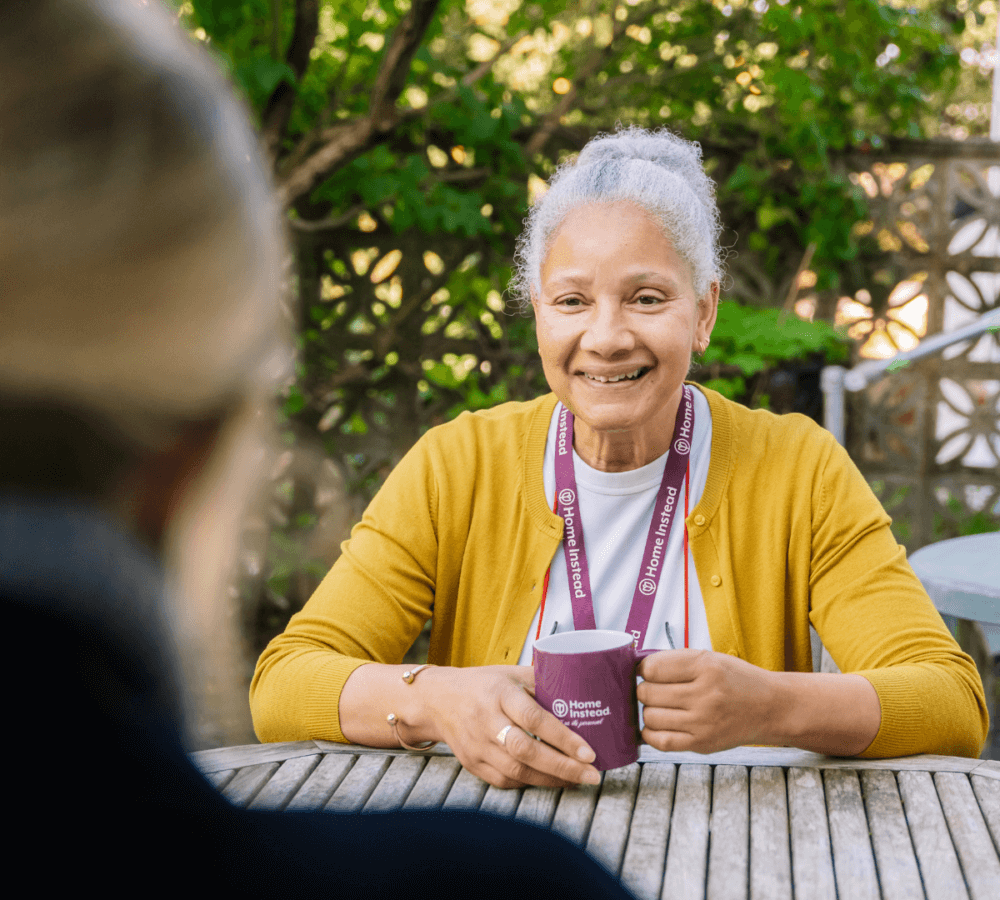 A smiling person with grey hair in a yellow cardigan holds a mug, sitting outdoors at a wooden table. - Home Instead