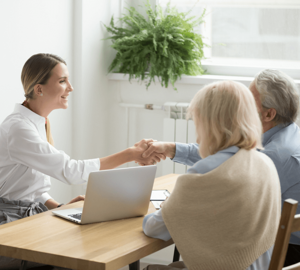 A smiling woman shakes hands with an elderly couple across a table with a laptop, next to a window with a plant. - Home Instead