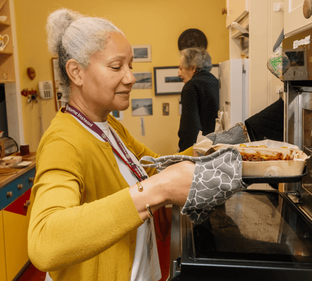 A woman in a yellow cardigan is taking a dish out of the oven in a home kitchen. Another person stands in the background. - Home Instead