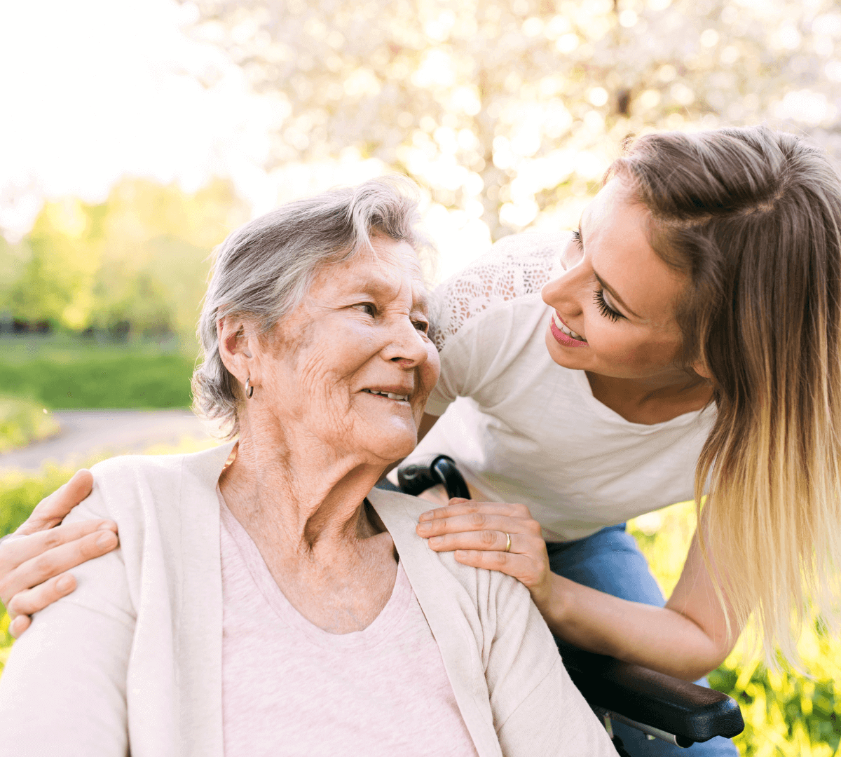 Younger woman smiles and leans over elderly woman in wheelchair outdoors, both look at each other affectionately. - Home Instead