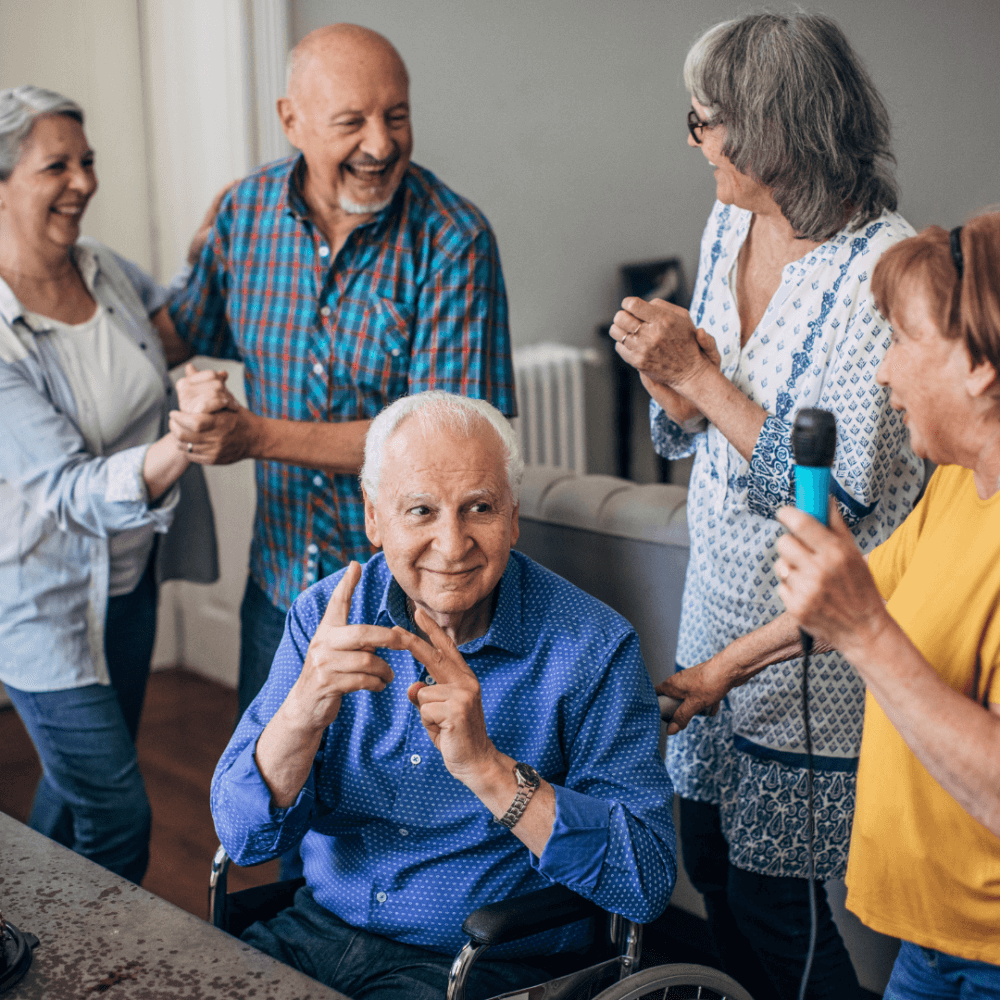 Group of seniors enjoying a lively moment together, laughing and singing, while one man sits in a wheelchair. - Home Instead