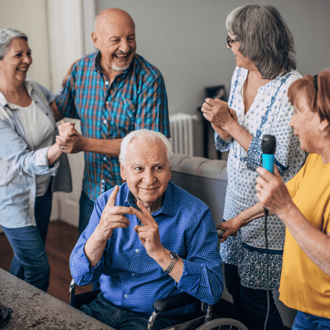 Group of seniors enjoying a lively moment together, laughing and singing, while one man sits in a wheelchair. - Home Instead