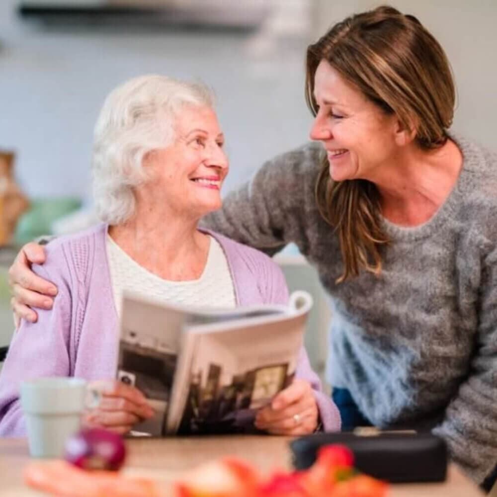 Older woman holding a magazine and sitting next to a younger woman in a cozy kitchen, both smiling at each other. - Home Instead