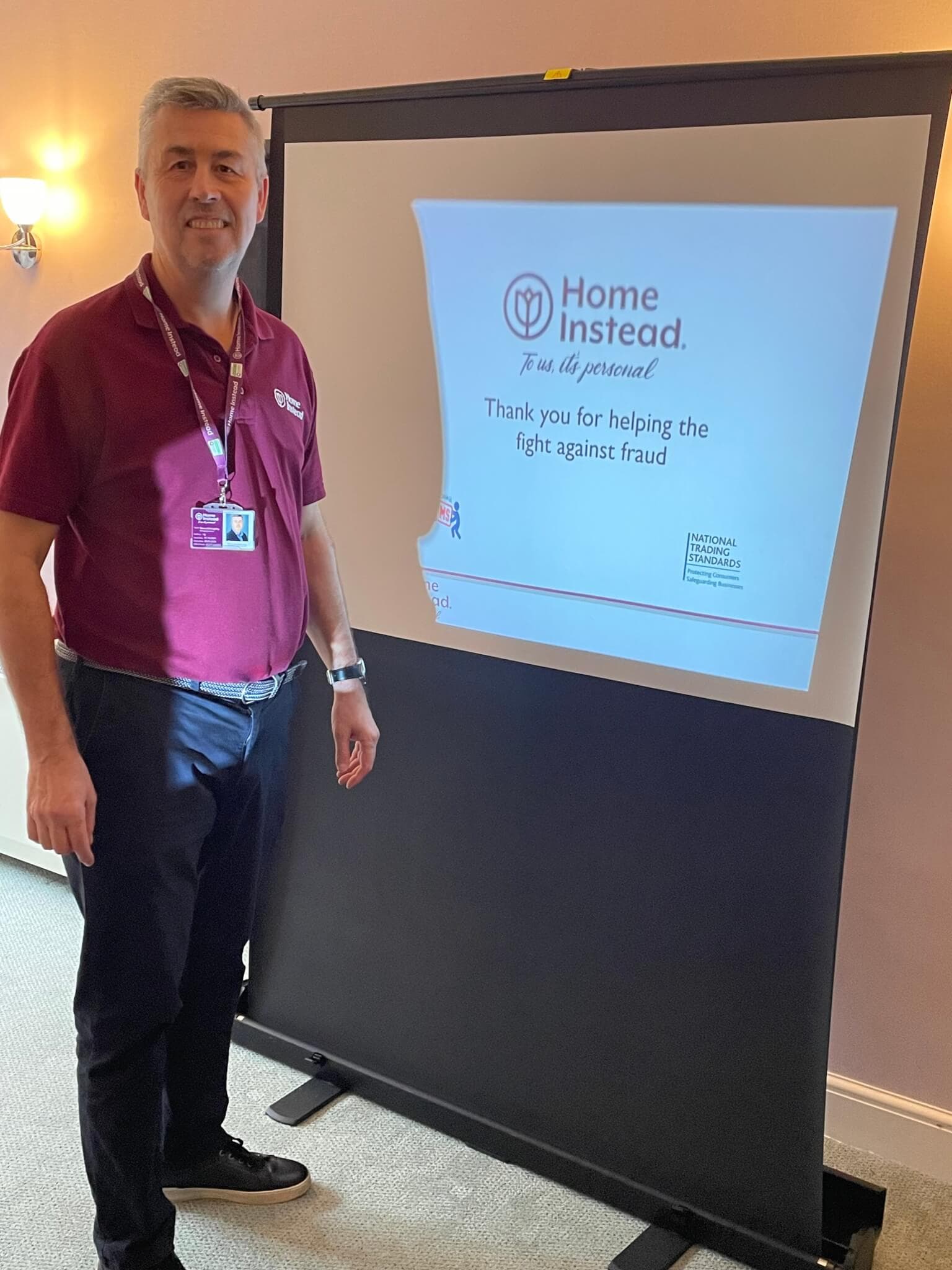 Man in a maroon shirt stands next to a flip chart with a presentation slide about fighting against fraud. - Home Instead