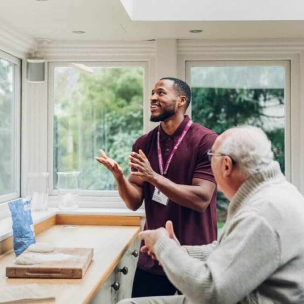 An elderly man and a younger man have a friendly conversation by a window in a bright room. - Home Instead