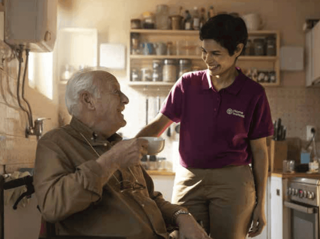 A caregiver in a uniform helps an elderly man in a wheelchair, holding a cup, in a warmly-lit kitchen. - Home Instead