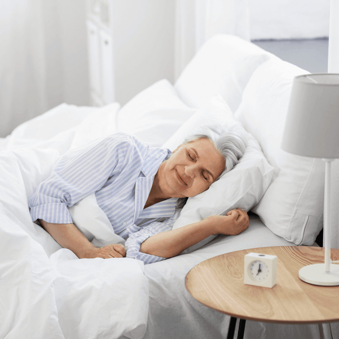 Elderly woman with gray hair sleeps peacefully in bed with white bedding, a nightstand, and a clock beside her. - Home Instead