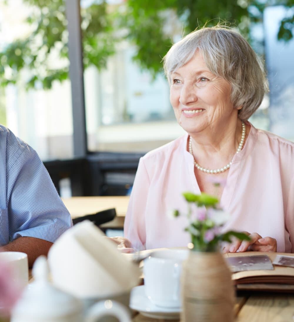 Elderly woman with gray hair smiles while seated at a table in a bright café, holding a book. - Home Instead