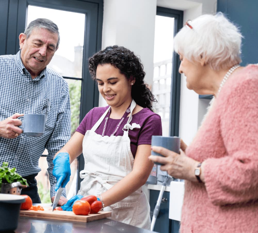 A young woman chops vegetables in a kitchen, while two seniors stand nearby with cups, chatting and smiling. - Home Instead
