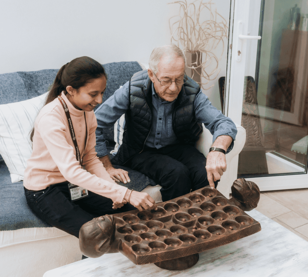An elderly man and a young girl sit on a couch playing a board game made of dark wood. - Home Instead Poole