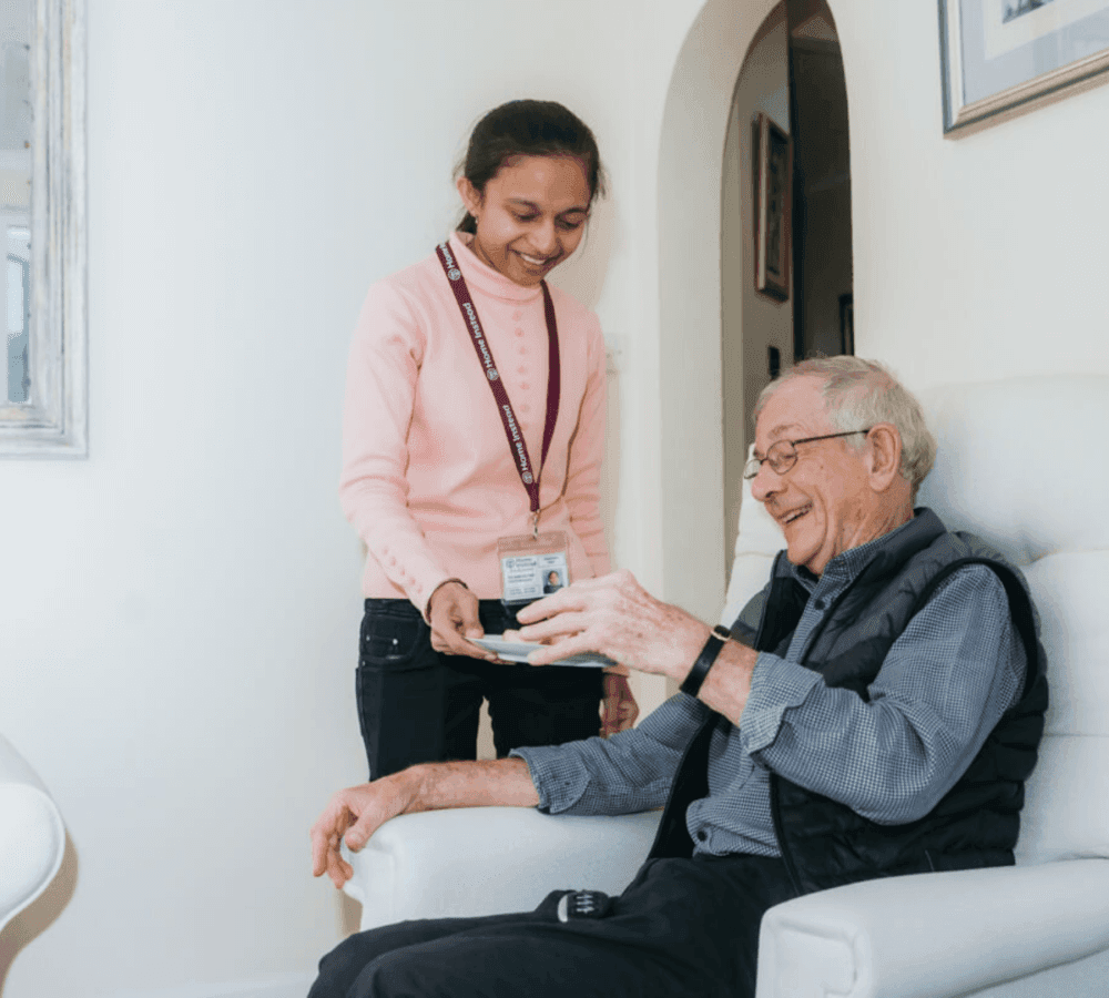A caregiver in a pink sweater hands an elderly man a drink as he sits comfortably in a white chair, smiling. - Home Instead Poole