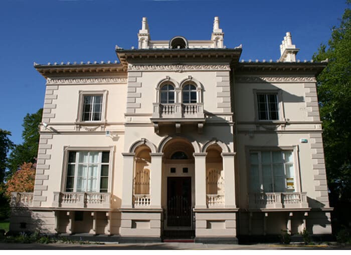 Large, historic two-story mansion with arched windows, cream-colored facade, and a small balcony on the second floor. - Home Instead