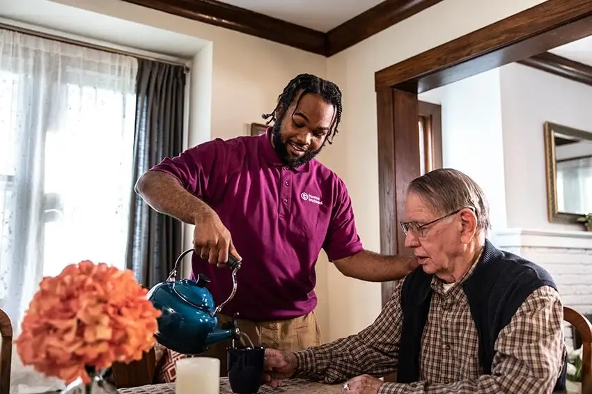 A caregiver in a purple shirt pours tea for an elderly man at a dining table with flowers in a bright cozy room. - Home Instead