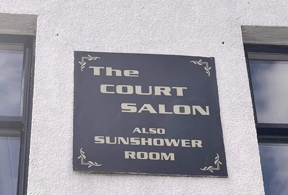 A sign on a white wall reads "The Court Salon Also Sunshower Room" between two windows. - Home Instead