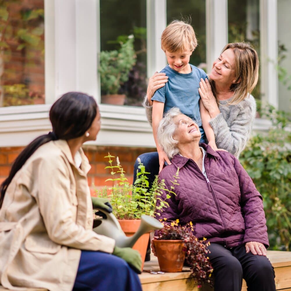 Three generations enjoying garden time; an elderly woman, a young boy, and two women smiling and talking. - Home Instead