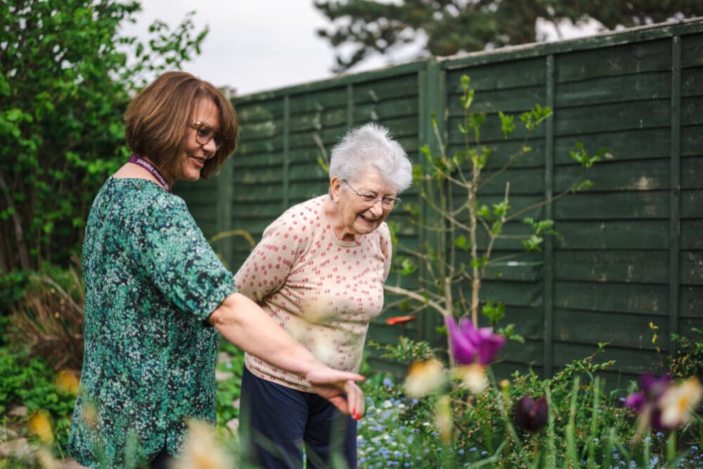 Two women, one elderly and one middle-aged, enjoying a garden with various blooming flowers beside a wooden fence. - Home Instead