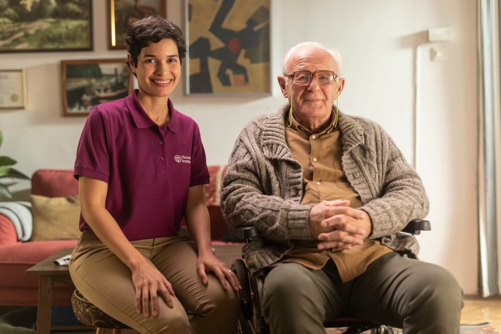A caregiver in a purple shirt sits beside an elderly man in a wheelchair, both smiling in a cozy room. - Home Instead