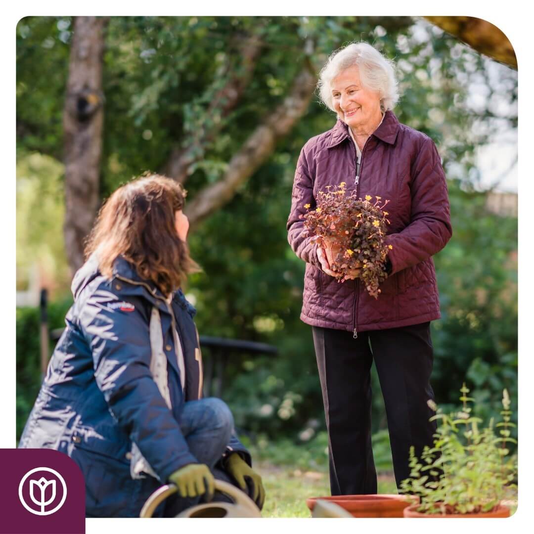 Two women gardening; one stands holding a plant, while the other kneels, both smiling. Trees in the background. - Home Instead
