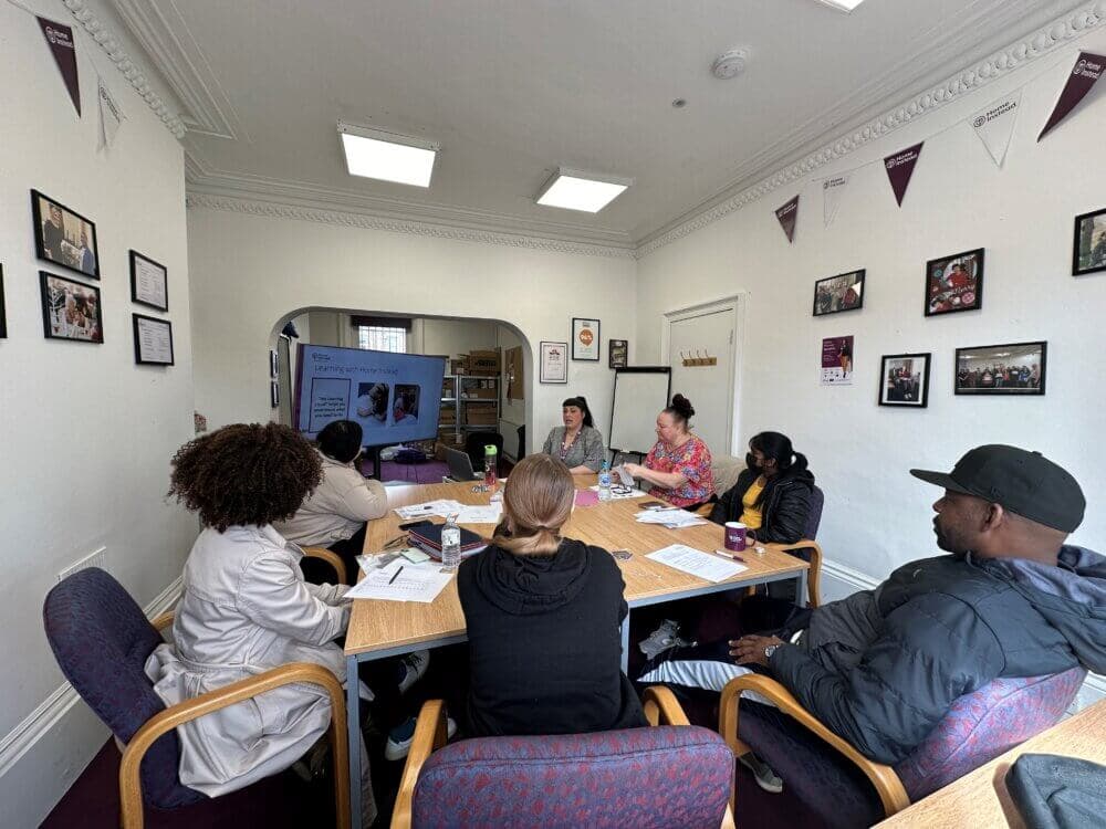 Six people sitting around a table in a meeting room, watching a presentation on a TV screen. - Home Instead
