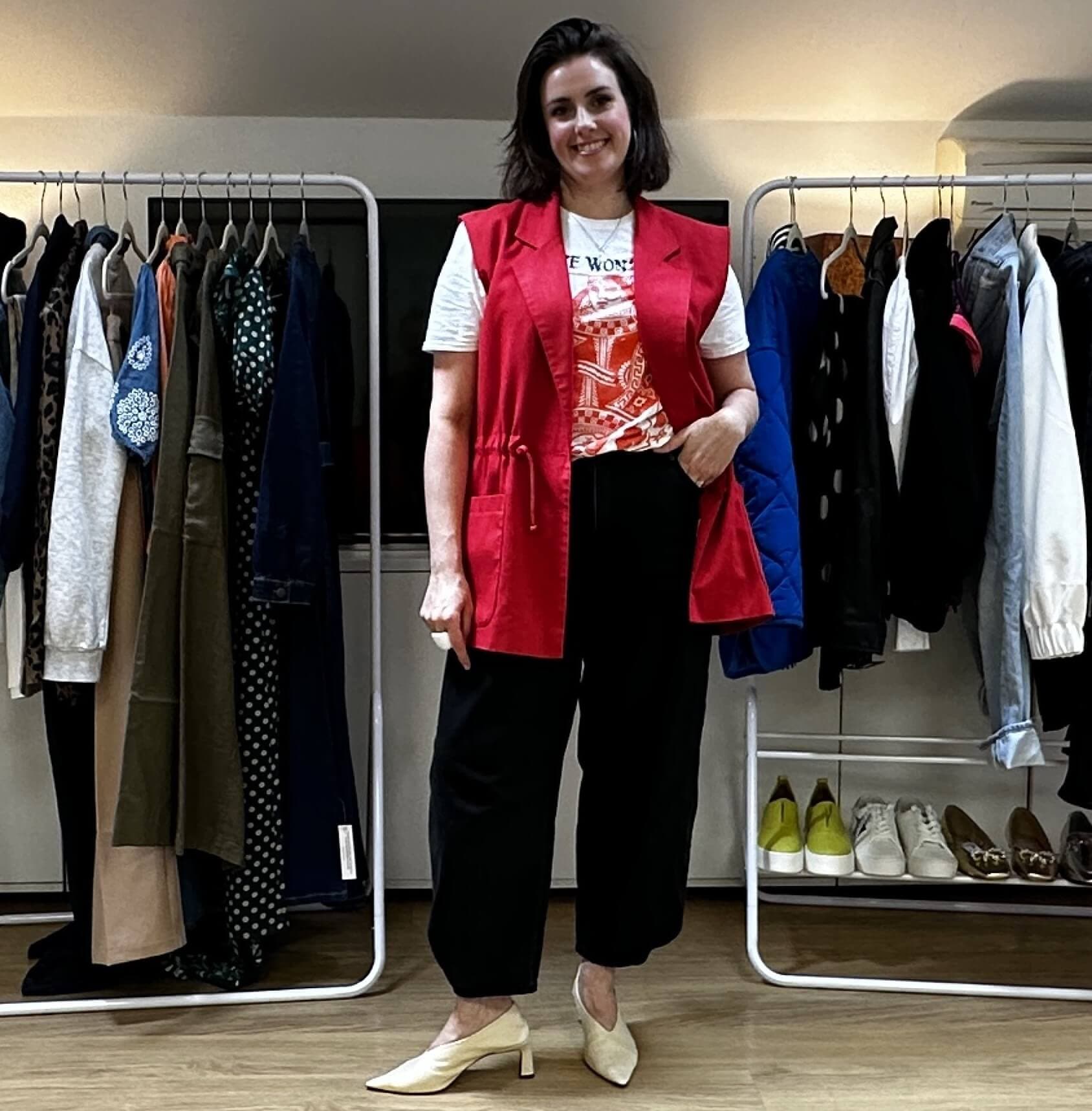 Smiling woman in a red vest and black pants stands between clothing racks in a room with hardwood floors. - Home Instead