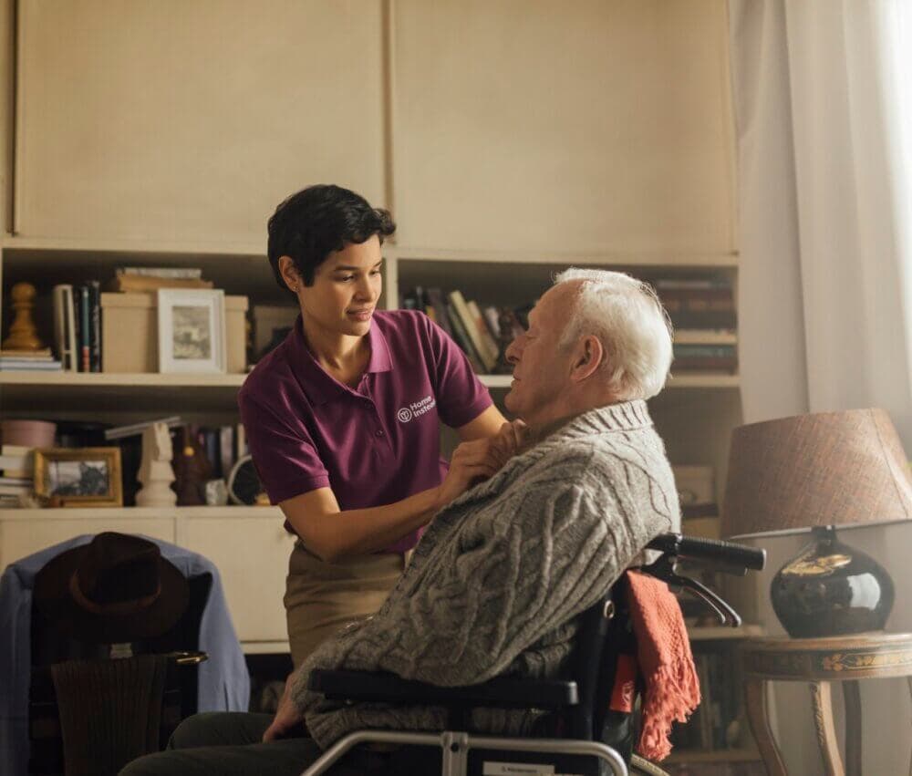 A caregiver in a purple shirt helps an elderly man in a wheelchair adjust his sweater in a cozy, book-filled room. - Home Instead