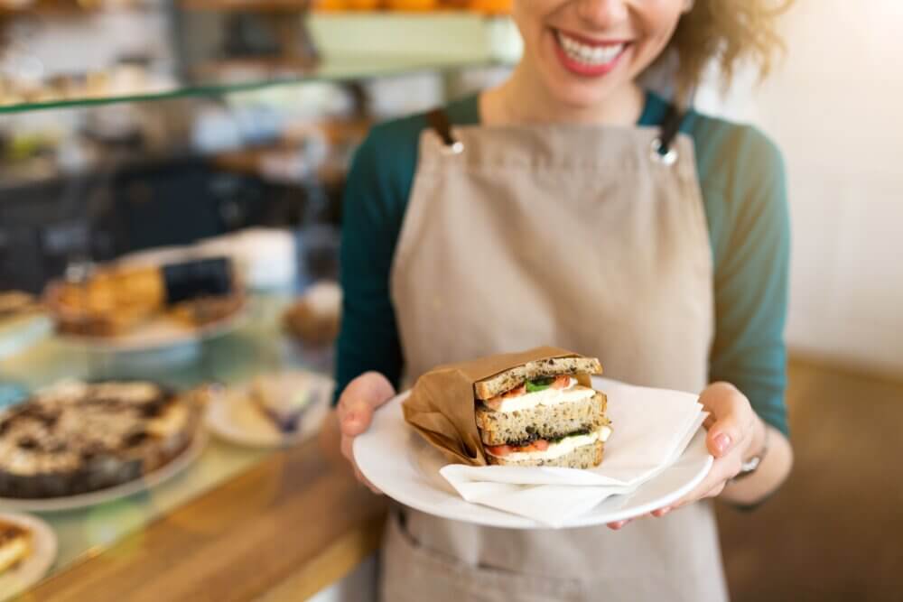 Smiling person holding a plate with a wrapped sandwich, standing in a café with pastries in the background. - Home Instead