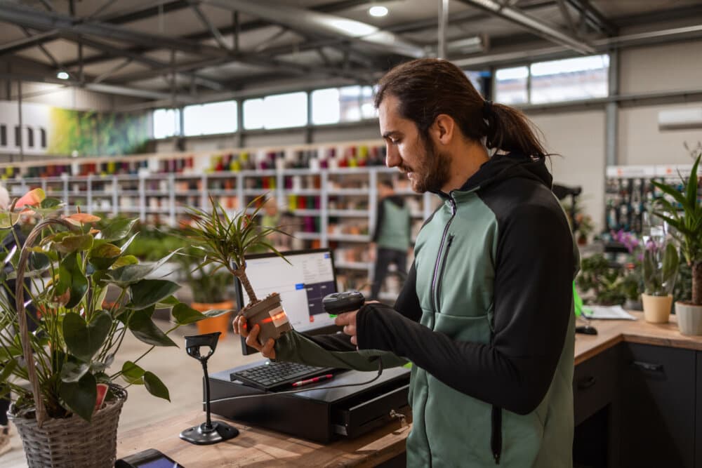 A man behind the cashier stand is scanning a price tag on a potted plant with a barcode reader. - Home Instead