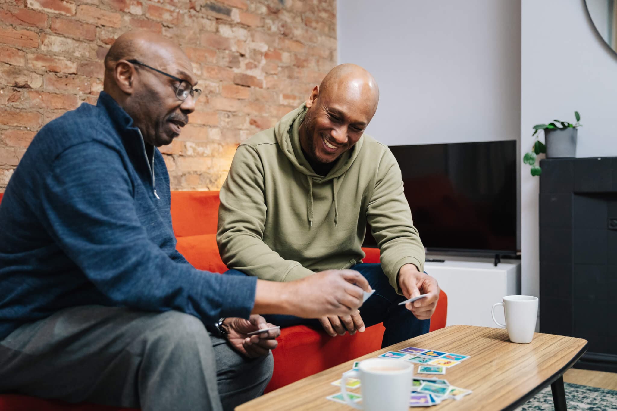 Two men sit on a couch, enjoying a card game and laughing, with mugs on the table. - Home Instead