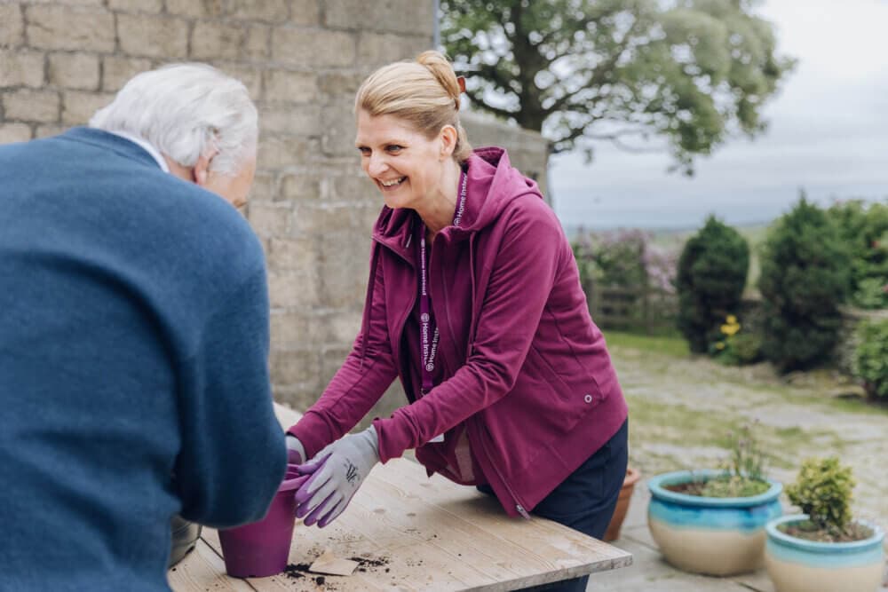 Two people at an outdoor table gardening together, laughing and enjoying the activity. - Home Instead