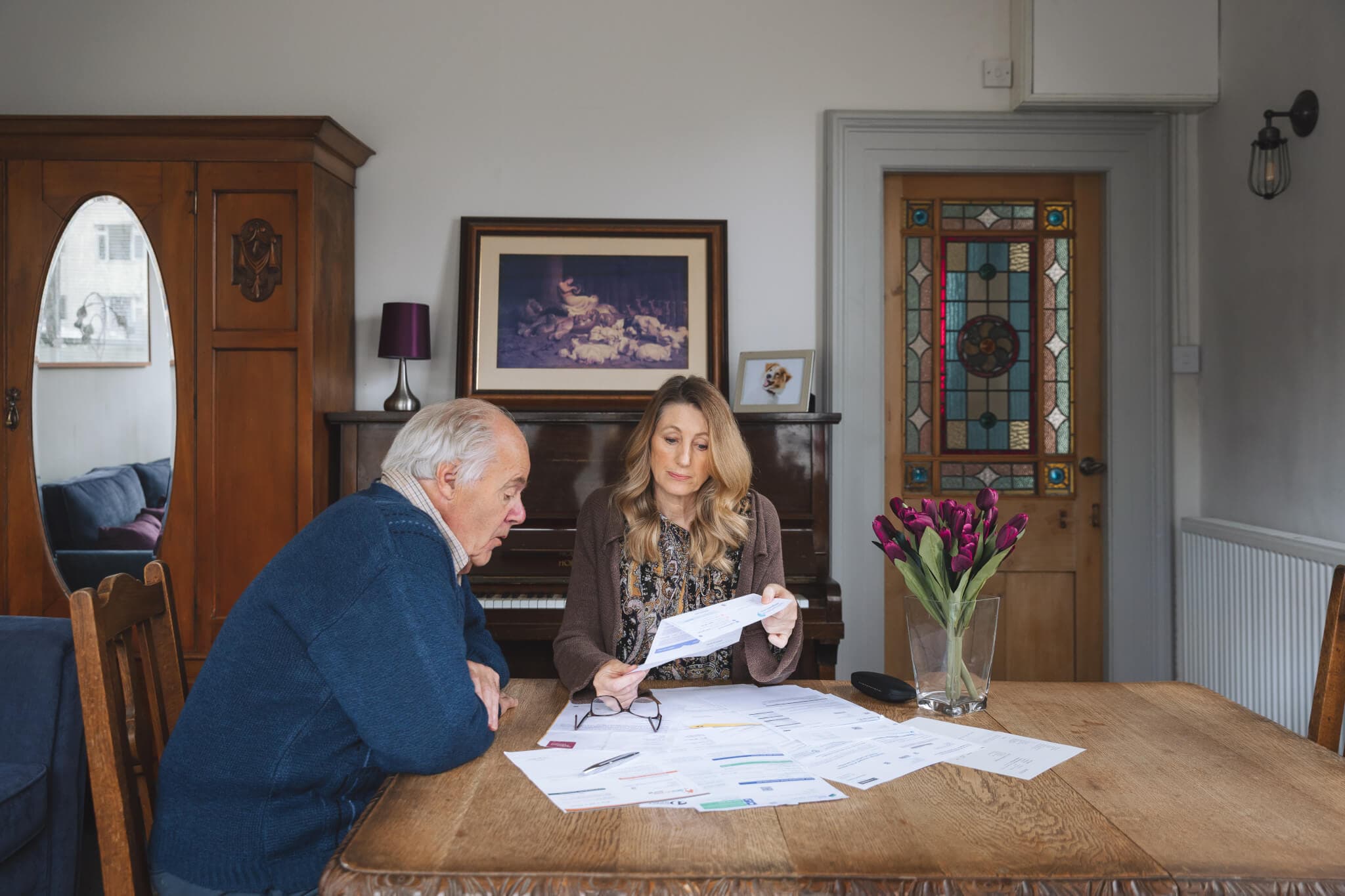 An elderly man and a woman review documents at a wooden table in a cozy room with stained glass door and flowers. - Home Instead