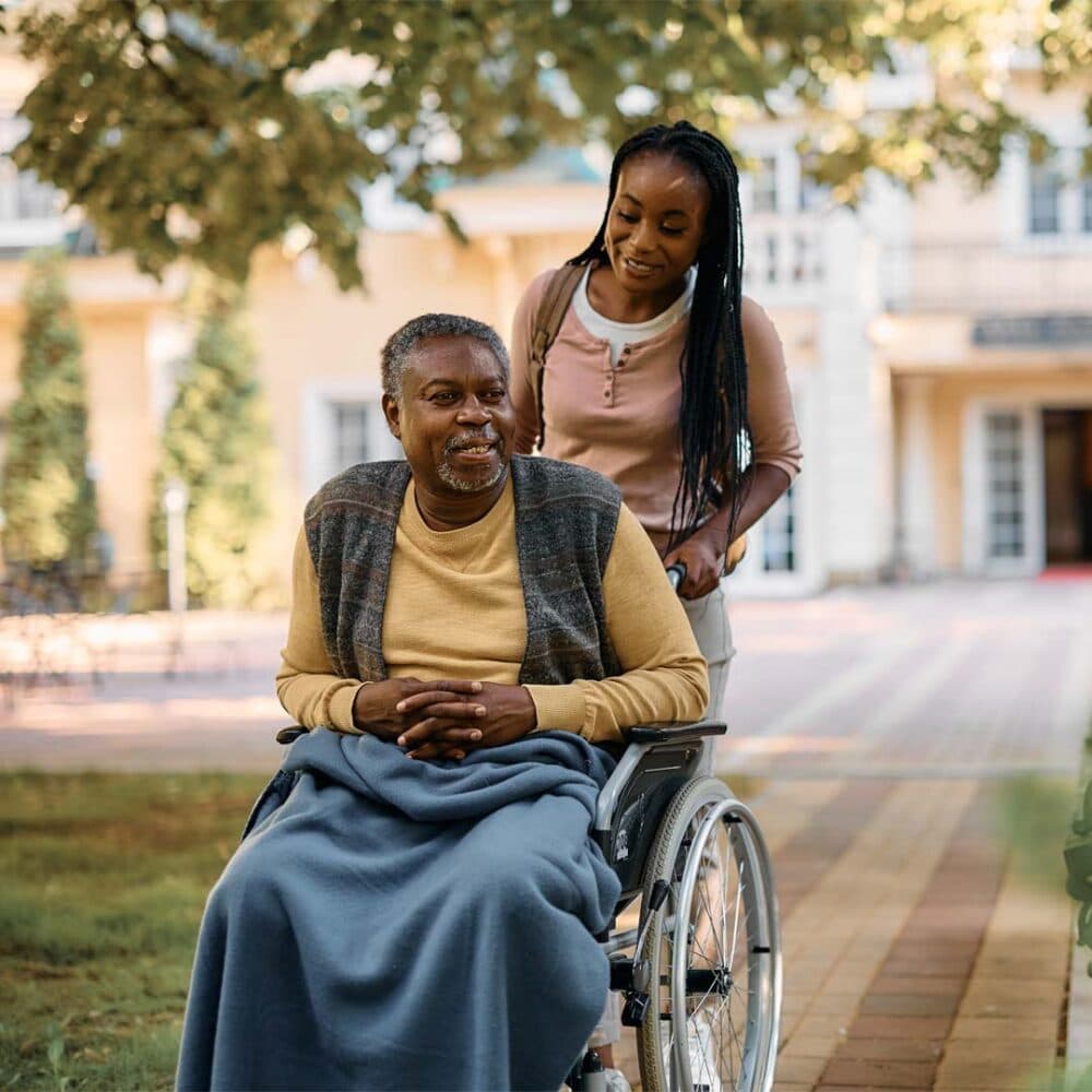 A woman pushes a man in a wheelchair outside, surrounded by trees and a building in the background, illustrating how outdoor activities contribute to preventing hospital admissions.