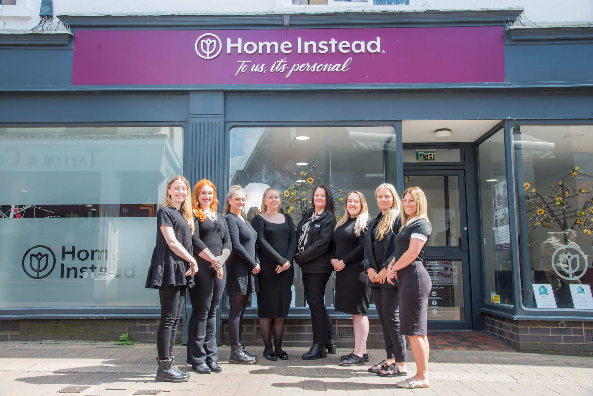 A group of nine women stands in front of a Home Instead office under a sign that reads, "To us, it's personal. - Home Instead