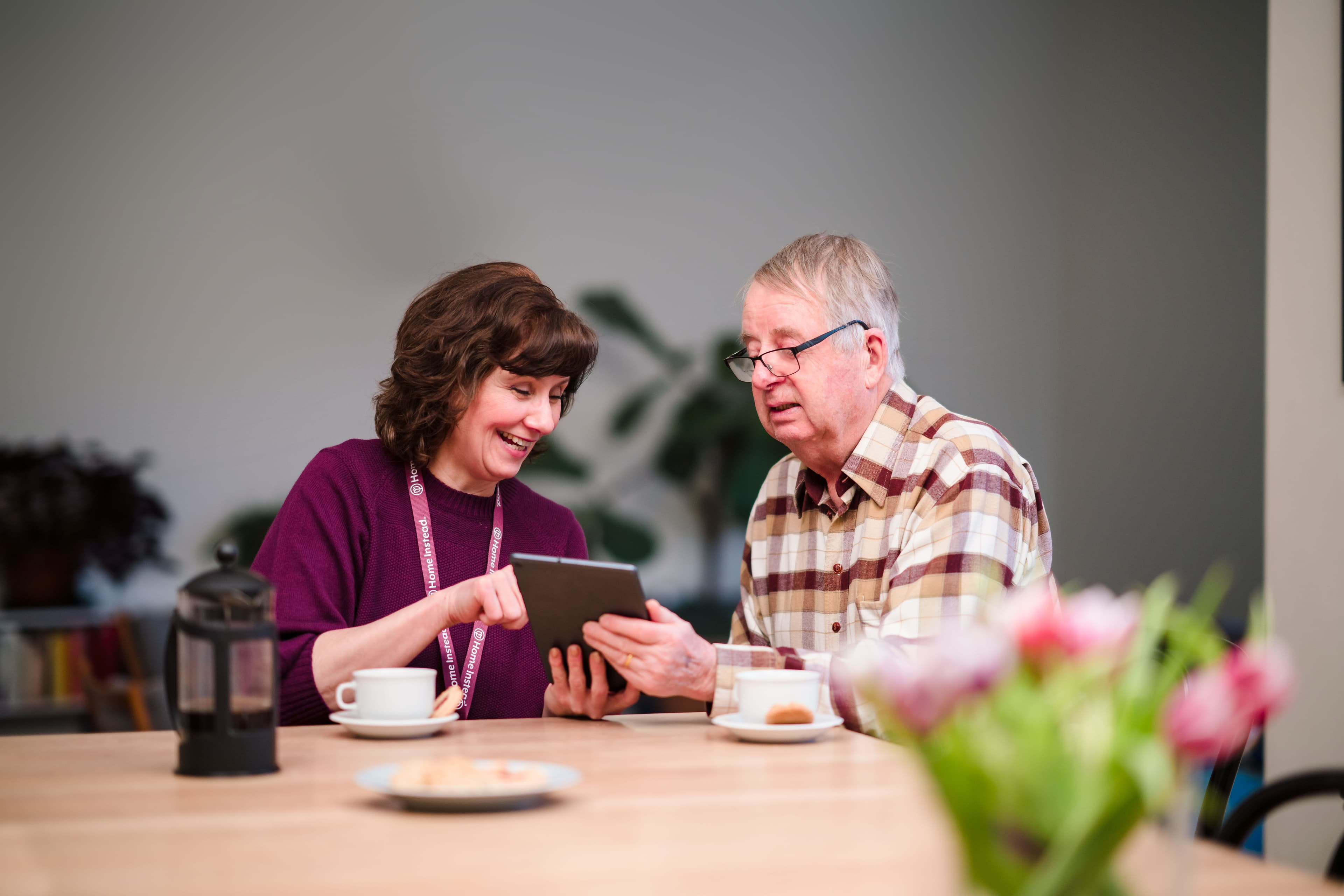 A woman helps an elderly man navigate a tablet at a table with coffee and flowers, both smiling and engaged. - Home Instead