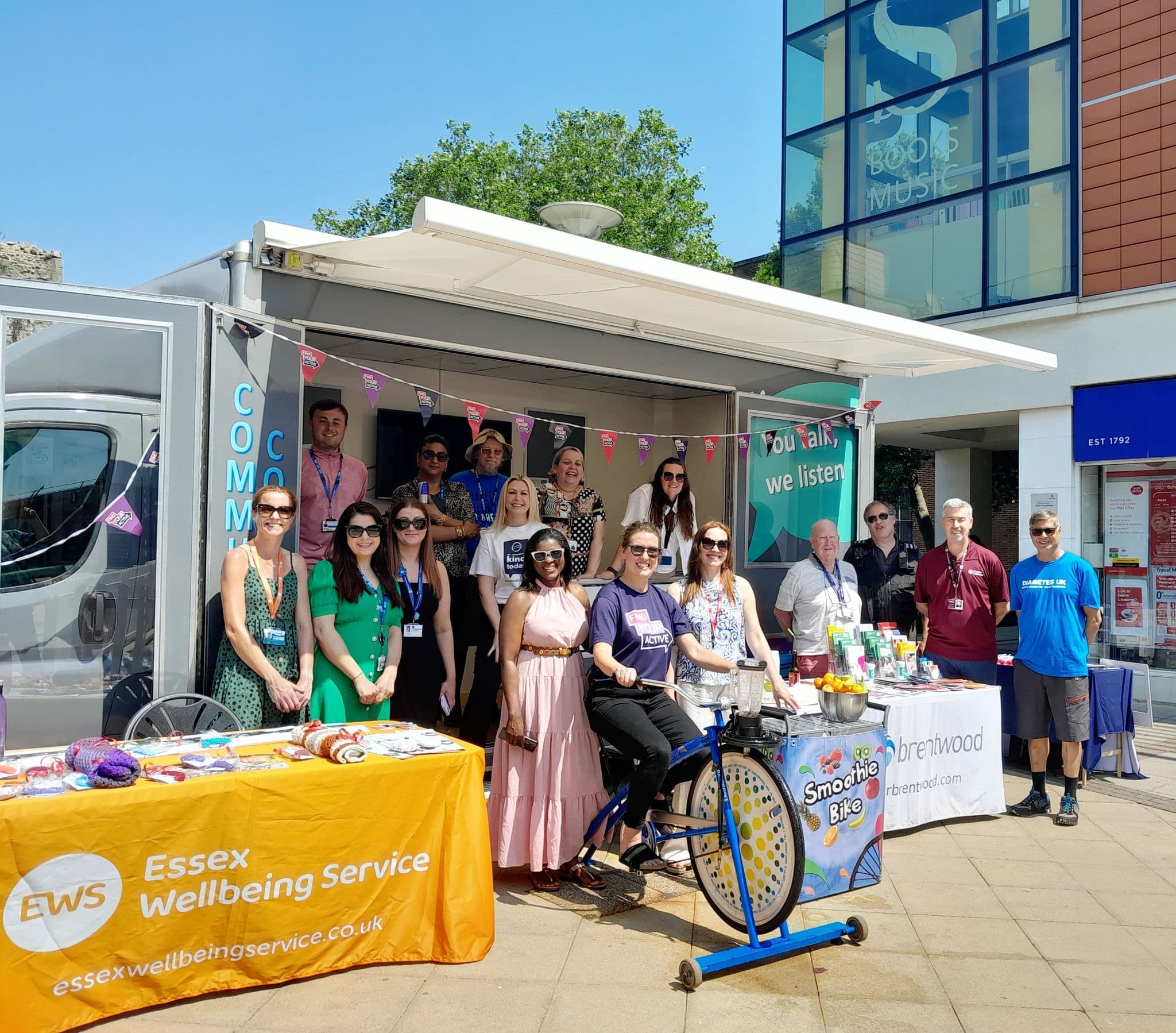 Group of people standing in front of stalls and a van at an outdoor event, with banners for Essex Wellbeing Service. - Home Instead