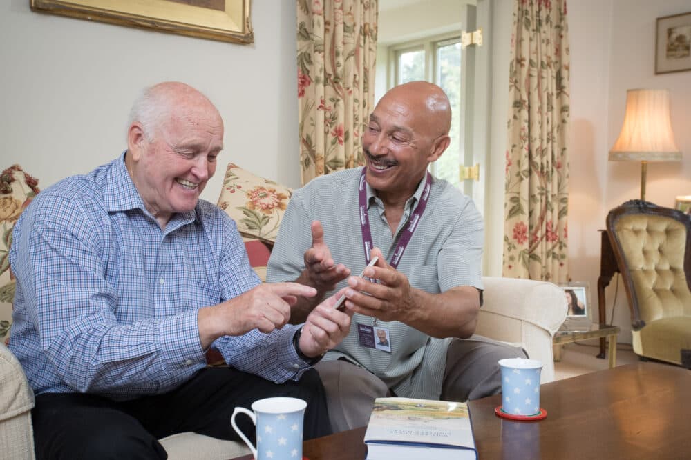 A caregiver and an elderly man sit together on a couch, smiling and pointing at something while sharing a joyful moment. - Home Instead