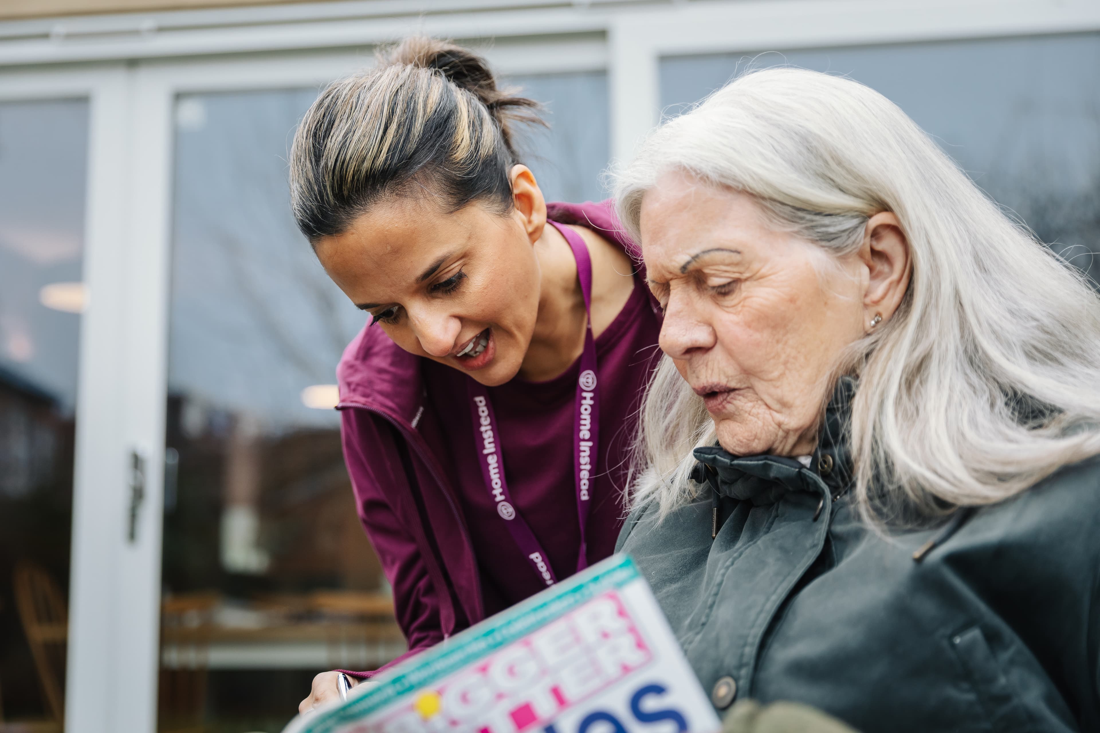 A caregiver with a badge helps an elderly woman read a magazine in front of a building with large windows. - Home Instead