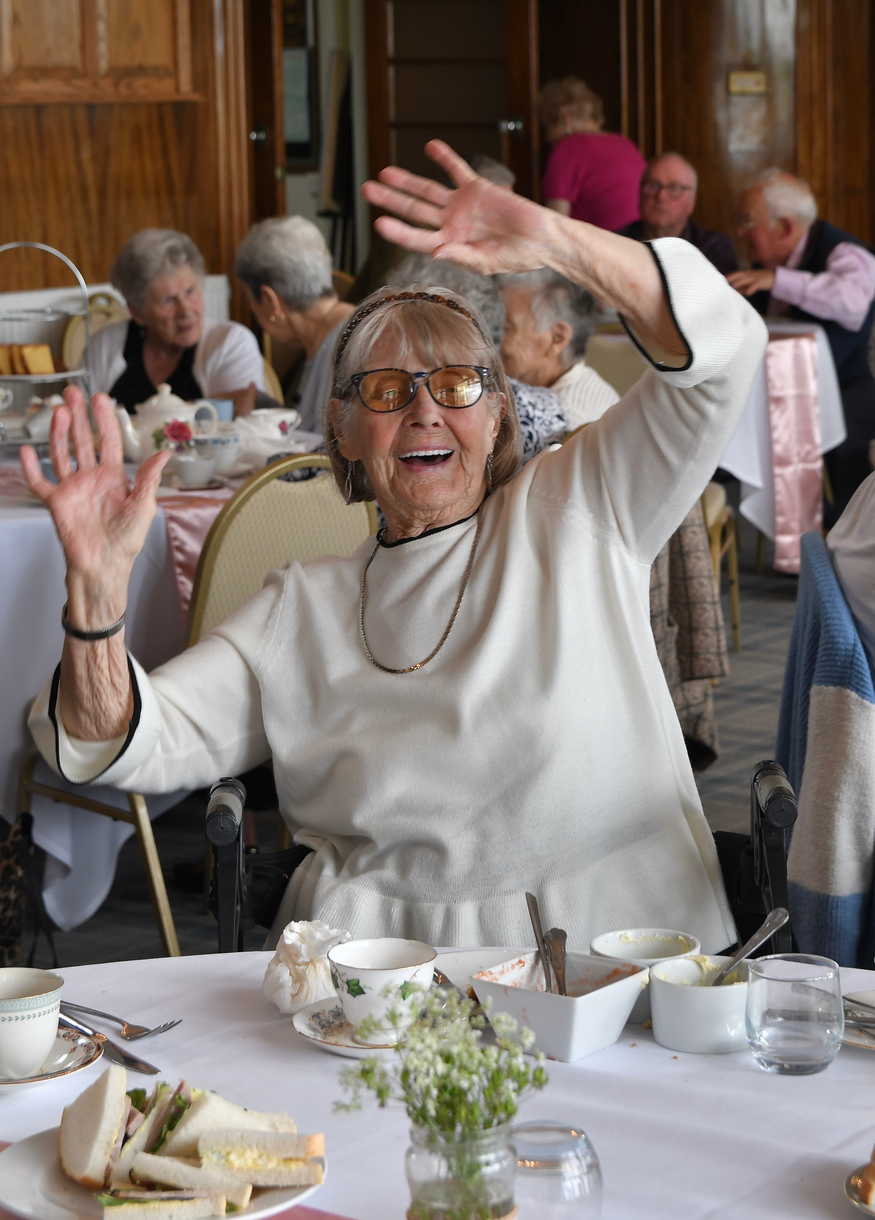 Elderly woman in white smiling and raising her hands at a lively tea party with others in the background. - Home Instead