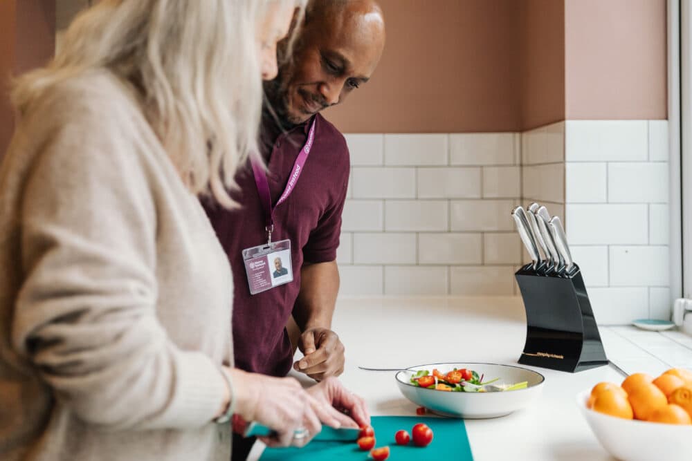 Two people in a kitchen, one chopping tomatoes on a cutting board, with a bowl of salad and a knife block in the background. - Home Instead